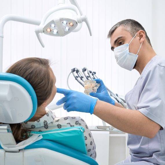 Dentist with blue gloves shows dental model to seated female patient in modern clinic with overhead light